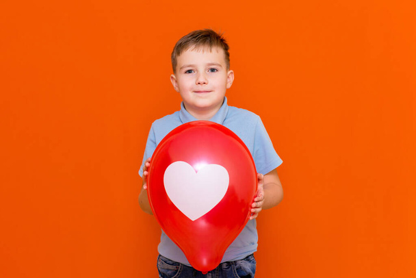 Close up Valentine's Day. Happy caucasian joyful young boy holding a big red balloon.Kid with a white drawn heart on balloon.Orange studio background. - Foto, afbeelding