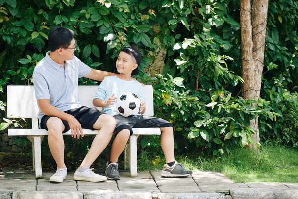 Vietnamese father and his preteen son resting on bench after playing soccer outdoors on Sunday - Photo, Image
