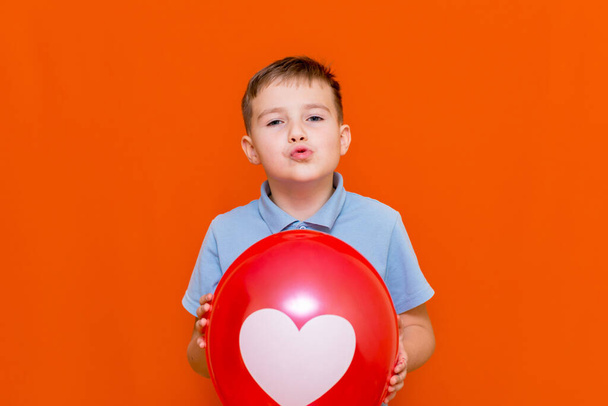 Close up Valentine's Day. Happy caucasian joyful young boy holding a big red balloon.Kid with a white drawn heart on balloon.Orange studio background. - Fotó, kép