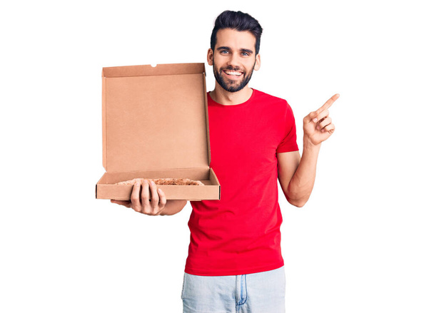 Young handsome man with beard holding delivery cardoboard with italian pizza smiling happy pointing with hand and finger to the side  - Foto, Imagen