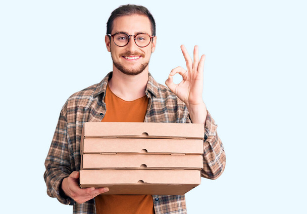 Молодий красивий кавказький чоловік тримає коробку для доставки піци робить знак з пальцями, посміхаючись дружній жест відмінний символ
  - Фото, зображення