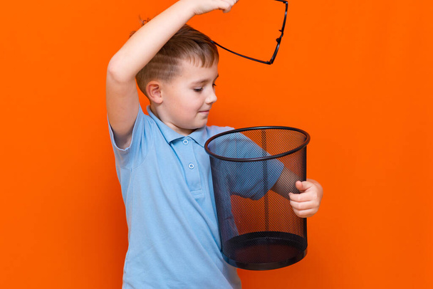 Вид сбоку Закрыть школьник в синей футболке бросает очки в мусорное ведро на фоне оранжевой студии - Фото, изображение