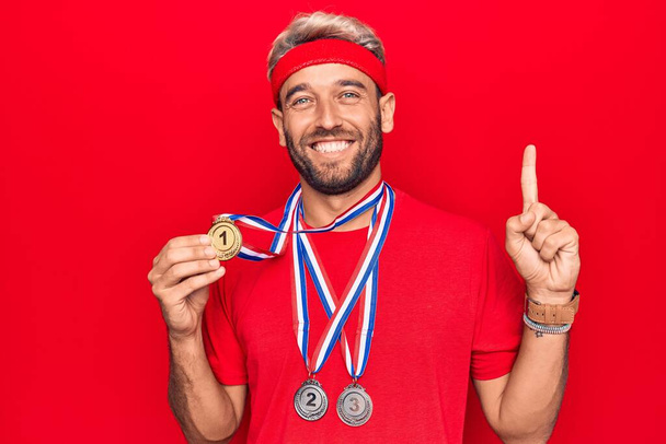 Mladý pohledný blonďatý sportovec s vousy vítěznými medailemi nad izolovaným červeným pozadím s úsměvem nápadem nebo otázkou ukazující prstem se šťastnou tváří, číslo jedna - Fotografie, Obrázek