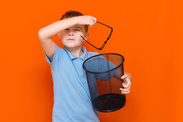 Close up the boy will shint in a blue t-shirt throw glasses in the bin on a orange studio background - Fotografie, Obrázek