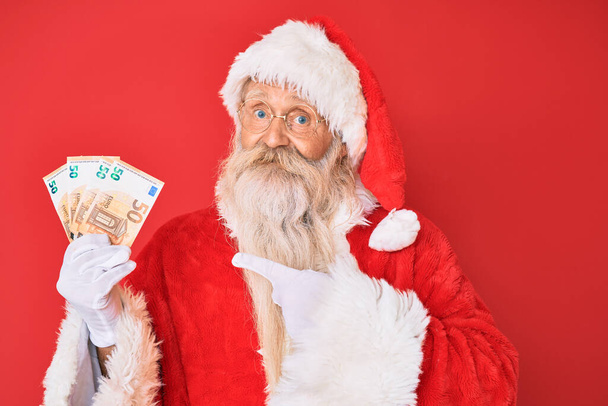 Stary staruszek w kostiumie Świętego Mikołaja trzymający euro uśmiechnięty szczęśliwy wskazując ręką i palcem  - Zdjęcie, obraz