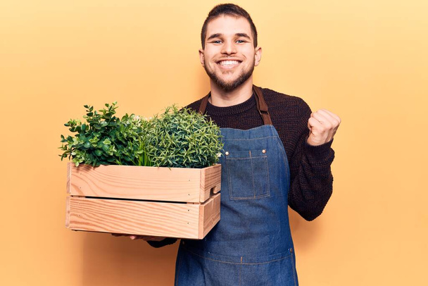 Young handsome man wearing gardener apron holding pant pot screaming proud, celebrating victory and success very excited with raised arm  - Foto, Bild