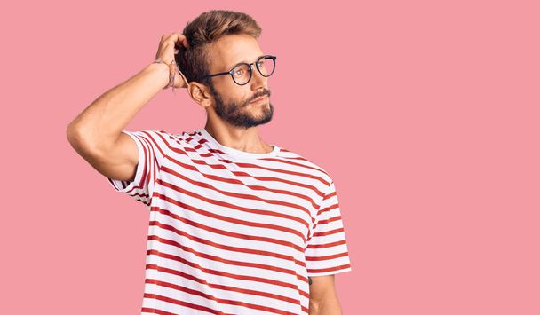 Handsome blond man with beard wearing casual clothes and glasses confuse and wondering about question. uncertain with doubt, thinking with hand on head. pensive concept.  - Photo, Image