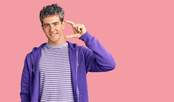 Fiatal jóképű férfi alkalmi lila pulóvert visel mosolygós és magabiztos gesztus kézzel csinál kis méretű jel ujjak keres, és a kamera. intézkedés koncepciója.  - Fotó, kép