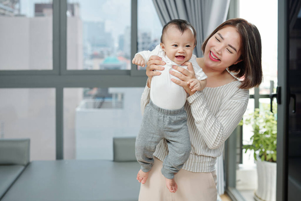 Χαρούμενη όμορφη Βιετναμέζα που κουβαλάει το μωρό της με βαμβακερό μπλουζάκι. - Φωτογραφία, εικόνα