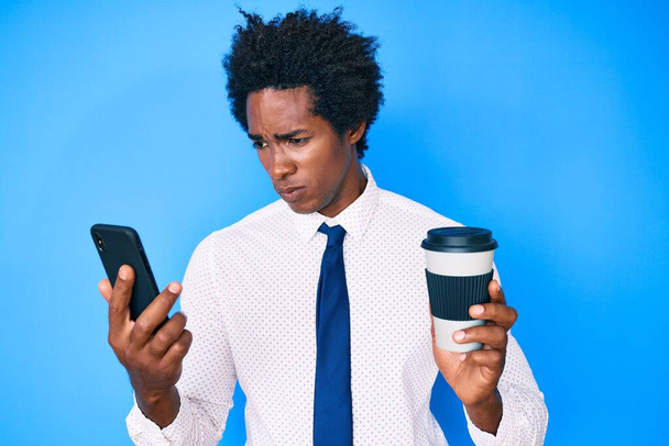 Hombre afroamericano guapo con cabello afro usando teléfono inteligente y bebiendo una taza de café escéptico y nervioso, frunciendo el ceño molesto debido a un problema. persona negativa.  - Foto, Imagen