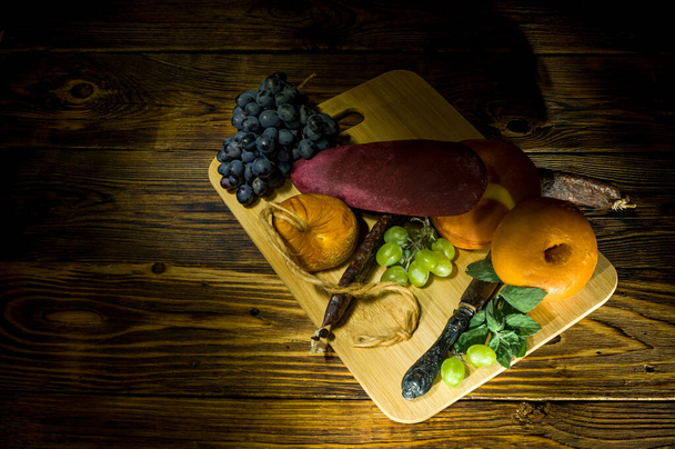 cheeses, sausage, sujuk, a bunch of blue grapes, a bunch of green grapes on a cutting board and a wooden table - 写真・画像