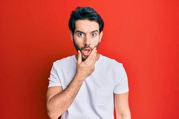 Young hispanic man wearing casual white tshirt looking fascinated with disbelief, surprise and amazed expression with hands on chin  - Foto, Imagem