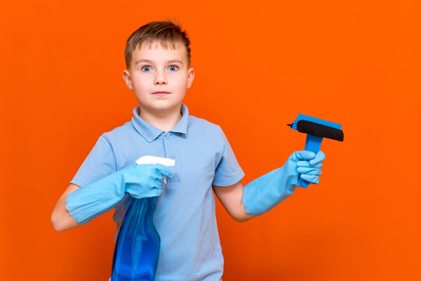 Concepto de limpieza. Retrato de estudio de niño apuesto colegial en guantes de látex limpiando vidrio con rociador deshumidificador de ventanas. Pared naranja. - Foto, imagen