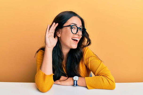 Mooie brunette jonge vrouw met een bril en casual kleding zitten op de tafel glimlachen met de hand over het oor luisteren naar een horen naar geruchten of roddels. doofheid.  - Foto, afbeelding