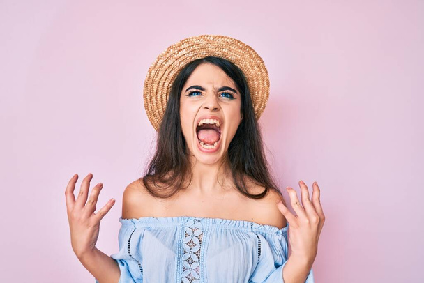 Brunette teenager girl wearing summer hat crazy and mad shouting and yelling with aggressive expression and arms raised. frustration concept.  - Photo, image