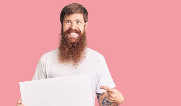 Schöner junger roter Mann mit langem Bart, der ein leeres Banner hält und glücklich lächelt und mit Hand und Finger zeigt  - Foto, Bild