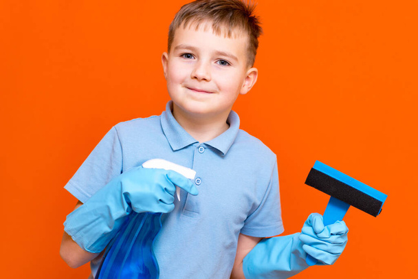 Housekeeping concept. Studio portrait of handsome kid child schoolboy in latex gloves cleaning glass with window dehumidifier sprayer. Orange wall. - 写真・画像