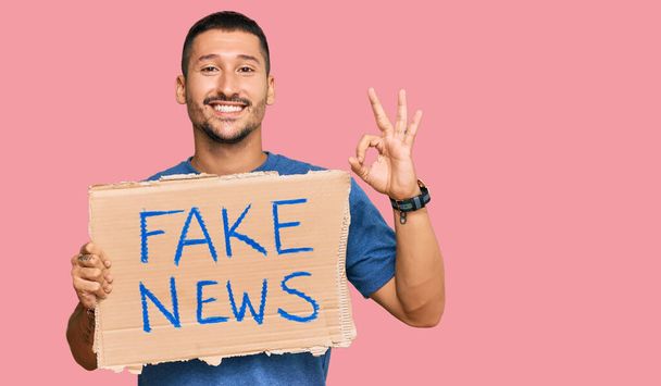 Handsome man with tattoos holding fake news banner doing ok sign with fingers, smiling friendly gesturing excellent symbol  - Photo, Image
