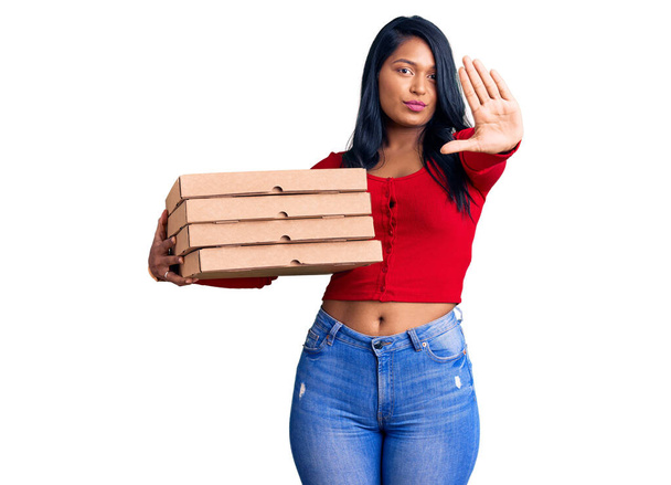 Hispanische Frau mit langen Haaren hält Lieferung Pizzaschachtel mit offener Hand tut Stoppschild mit ernstem und selbstbewusstem Gesichtsausdruck, Verteidigungsgeste  - Foto, Bild