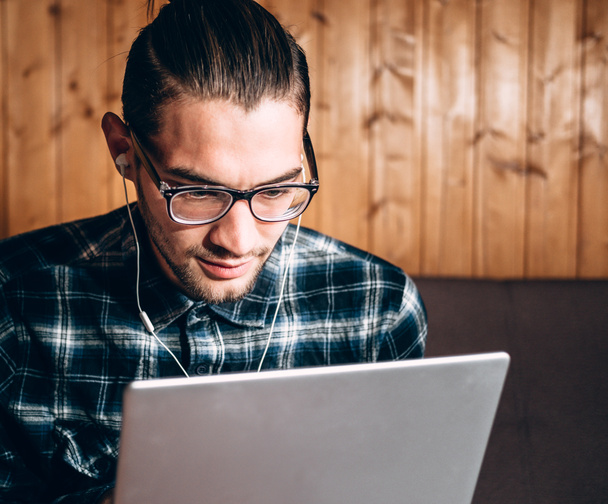 A handsome young freelancer in a blue shirt and glasses with diopters works on a silver laptop at home. Remote work during the quarantine. - Foto, afbeelding