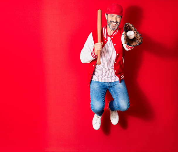 Ein gutaussehender Mann mittleren Alters in sportlicher Kleidung lächelt glücklich. Springen mit einem Lächeln auf dem Gesicht Baseball spielen mit Schläger, Ball und Handschuh über isolierten roten Hintergrund - Foto, Bild