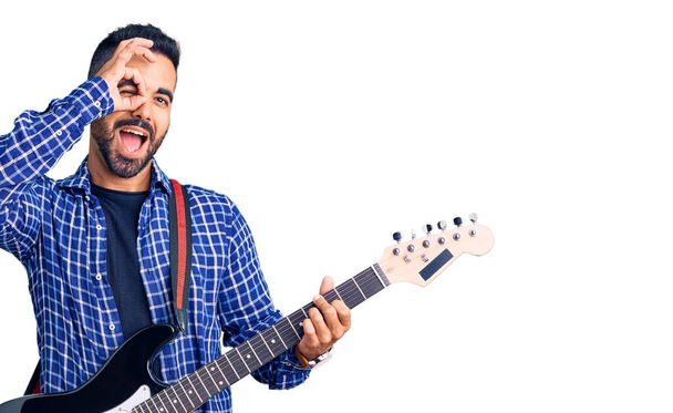 Joven hispano tocando la guitarra eléctrica sonriendo feliz haciendo ok signo con la mano en el ojo mirando a través de los dedos  - Foto, imagen