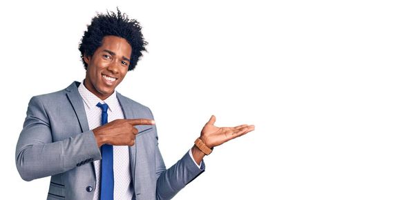 Нахабний африканський чоловік з афроамериканською зачіскою в діловій куртці, вражений і посміхаючись до камери, представляючи рукою і вказуючи пальцем..  - Фото, зображення