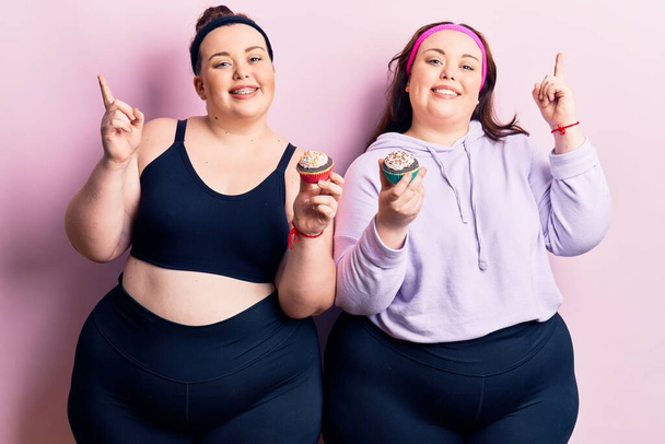 Joven más gemelos de tamaño usando ropa deportiva sosteniendo cupcake sonriendo feliz señalando con la mano y el dedo a un lado  - Foto, Imagen