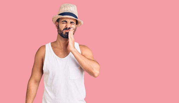 Młody Hiszpan noszący luźny letni kapelusz dotykający ust ręką z bolesnym wyrazem twarzy z powodu bólu zęba lub choroby zębów. dentysta  - Zdjęcie, obraz