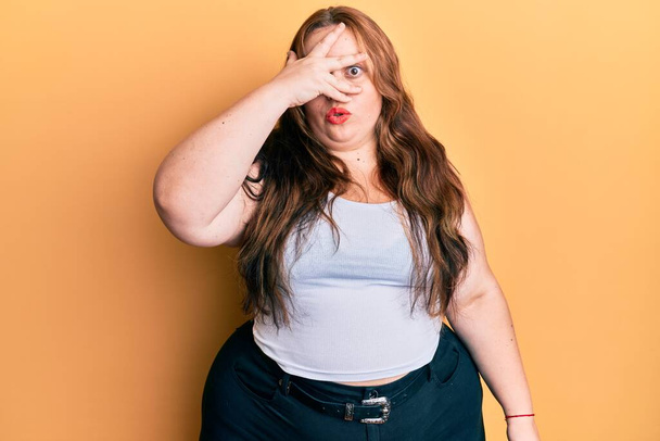 Plus size caucasian young woman wearing casual clothes peeking in shock covering face and eyes with hand, looking through fingers afraid  - Photo, Image
