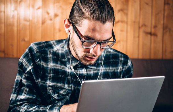 Un apuesto joven freelancer con camisa azul y gafas con dioptrías trabaja en una laptop plateada en casa. Trabajo remoto durante la cuarentena. - Foto, Imagen