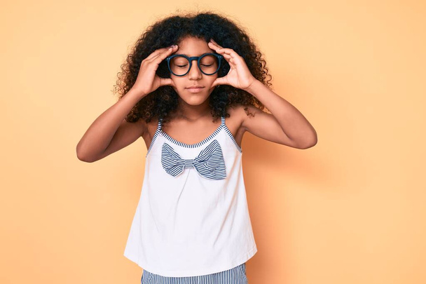 Afro-Amerikaans kind met krullend haar in casual kleding en bril met de hand op het hoofd, hoofdpijn omdat stress. lijden aan migraine.  - Foto, afbeelding