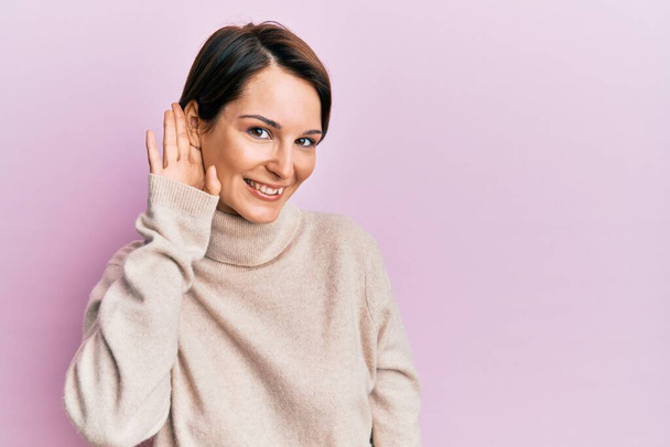 短い髪をした若いブルネットの女性は、噂やゴシップに耳を傾ける耳の上に手で笑みを浮かべてカジュアルな冬のセーターを着ていた。聴覚障害の概念.  - 写真・画像