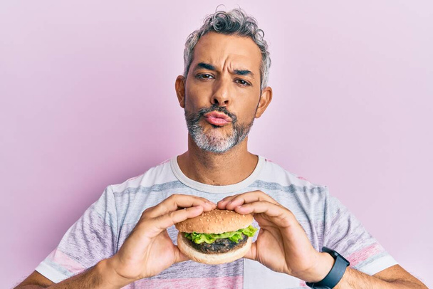 Middle age grey-haired man eating a tasty classic burger looking at the camera blowing a kiss being lovely and sexy. love expression.  - Fotó, kép