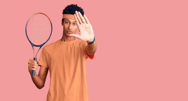 Молодий американець грає в теніс, тримаючи ракетку з відкритою рукою, роблячи знак "Стоп" з серйозним і впевненим виразом, захисний жест  - Фото, зображення