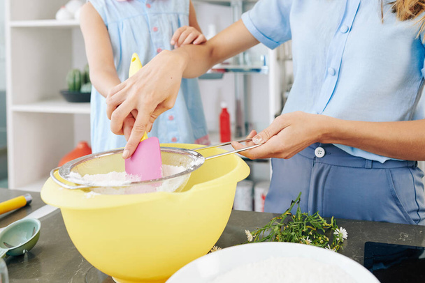Közelkép az anyáról, amint megmutatja a kiskorú lányának, hogyan kell lisztet szitálni, amikor tésztát csinálnak. - Fotó, kép