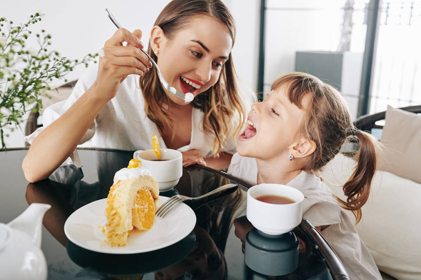 Cheerful mother giving piece of tasty homemade cake to little daughter when they are drinking tea together - Foto, Imagem