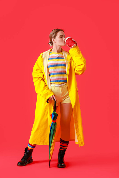 Beautiful young woman in raincoat, with coffee and umbrella on color background - Foto, Imagen