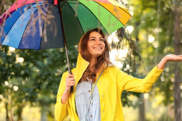 Όμορφη νεαρή γυναίκα με ομπρέλα φορώντας αδιάβροχο στο πάρκο - Φωτογραφία, εικόνα