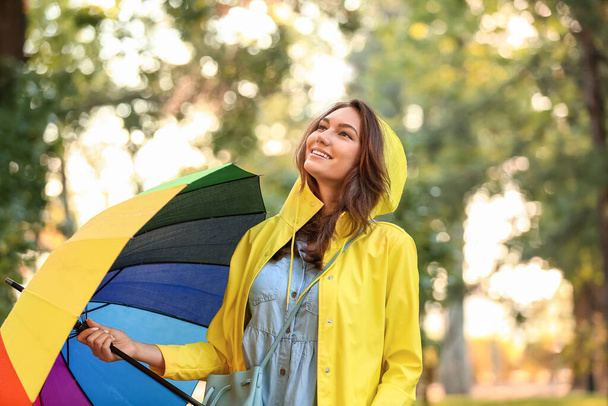 Όμορφη νεαρή γυναίκα με ομπρέλα φορώντας αδιάβροχο στο πάρκο - Φωτογραφία, εικόνα