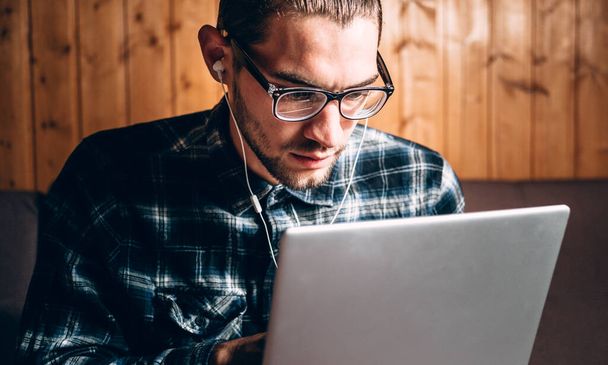 Un apuesto joven freelancer con camisa azul y gafas con dioptrías trabaja en una laptop plateada en casa. Trabajo remoto durante la cuarentena. - Foto, imagen