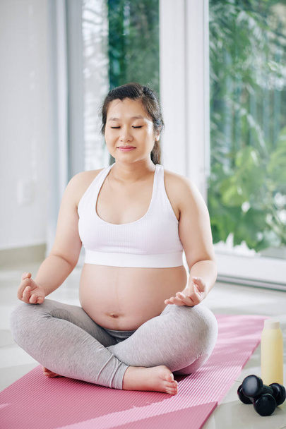 Mosolygó fiatal terhes vietnami nő ül lótuszpozícióban, csukott szemmel, és ezzel légzési gyakorlat - Fotó, kép