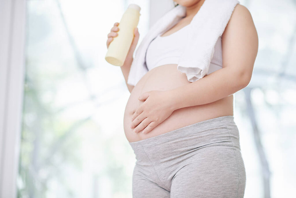 Oříznutý obraz těhotné ženy v obilí top a legíny pitné vody a dotýkání se jejího břicha po cvičení jógy - Fotografie, Obrázek