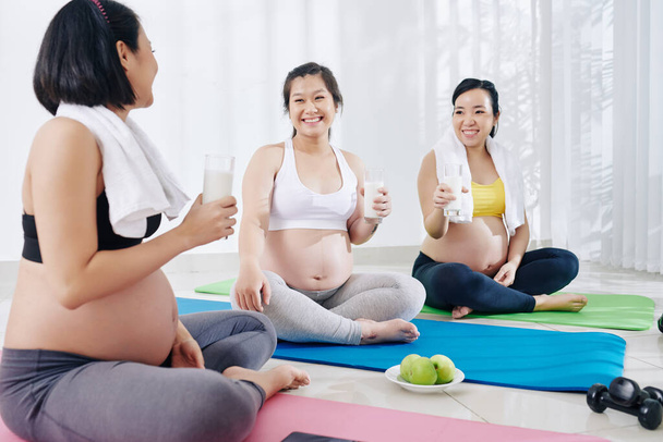 Gruppe fröhlich schwangerer asiatischer Frauen trinkt Joghurt und isst grüne Äpfel, wenn sie nach dem gemeinsamen Training über Mutterschaft sprechen - Foto, Bild