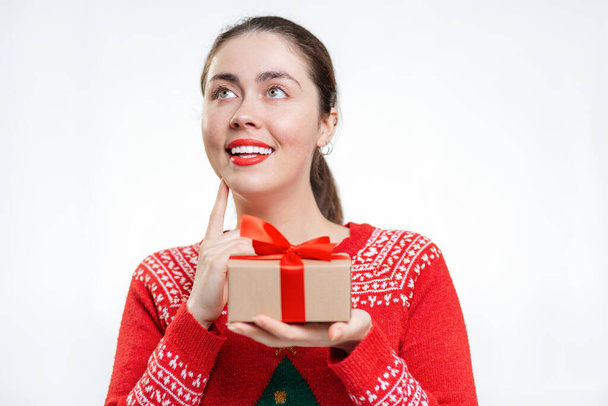 Portrait of a young smiling woman in a new year's sweater, holding a gift and making a wish. White background. Copy space. The concept of Christmas shopping. - Photo, Image