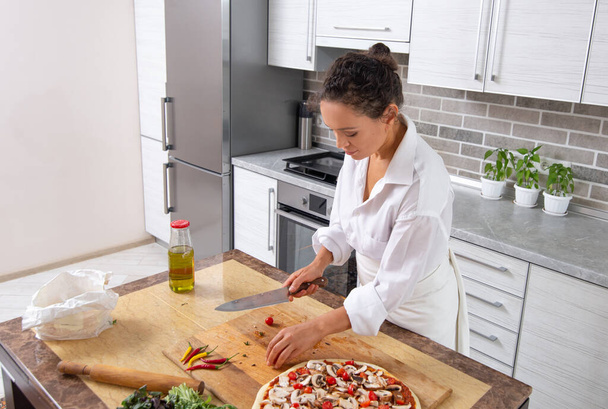 Die junge Köchin in der Küche schneidet Kirschtomaten für Pizza. - Foto, Bild