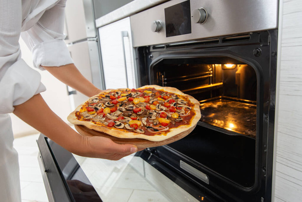 La giovane chef tira fuori una pizza cotta dal forno su una tavola di legno rotonda. - Foto, immagini