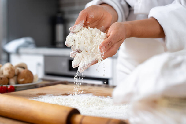 Close-up on the hands of a cook with a handful of white flour. Part of the flour spills onto the board on a table. On the table is a wooden board with scattered flour. - Photo, Image