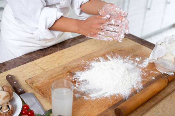 Detailní záběr na ruce kuchaře. Kuchař drtí mouku z jeho rukou na stůl. Na stole je dřevěná deska s roztroušenou moukou. Boční pohled. - Fotografie, Obrázek