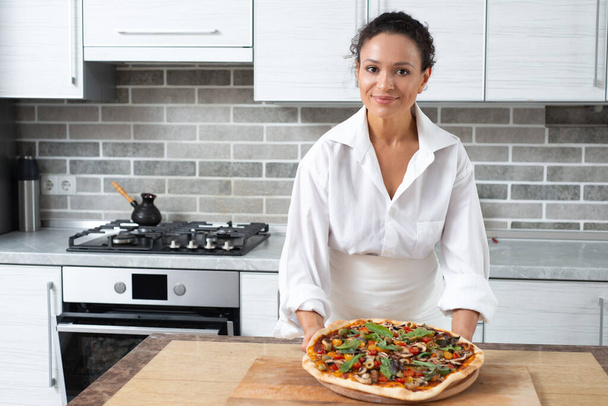 Genç kadın aşçı bize bakarken hazır pizza gösterisi sunuyor. Vejetaryen yemeği. - Fotoğraf, Görsel